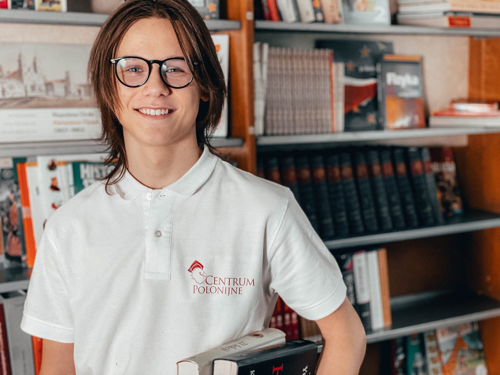 Усміхнений учень в окулярах і білому поло Centrum Polonijne з книжками біля полиць із польською літературою