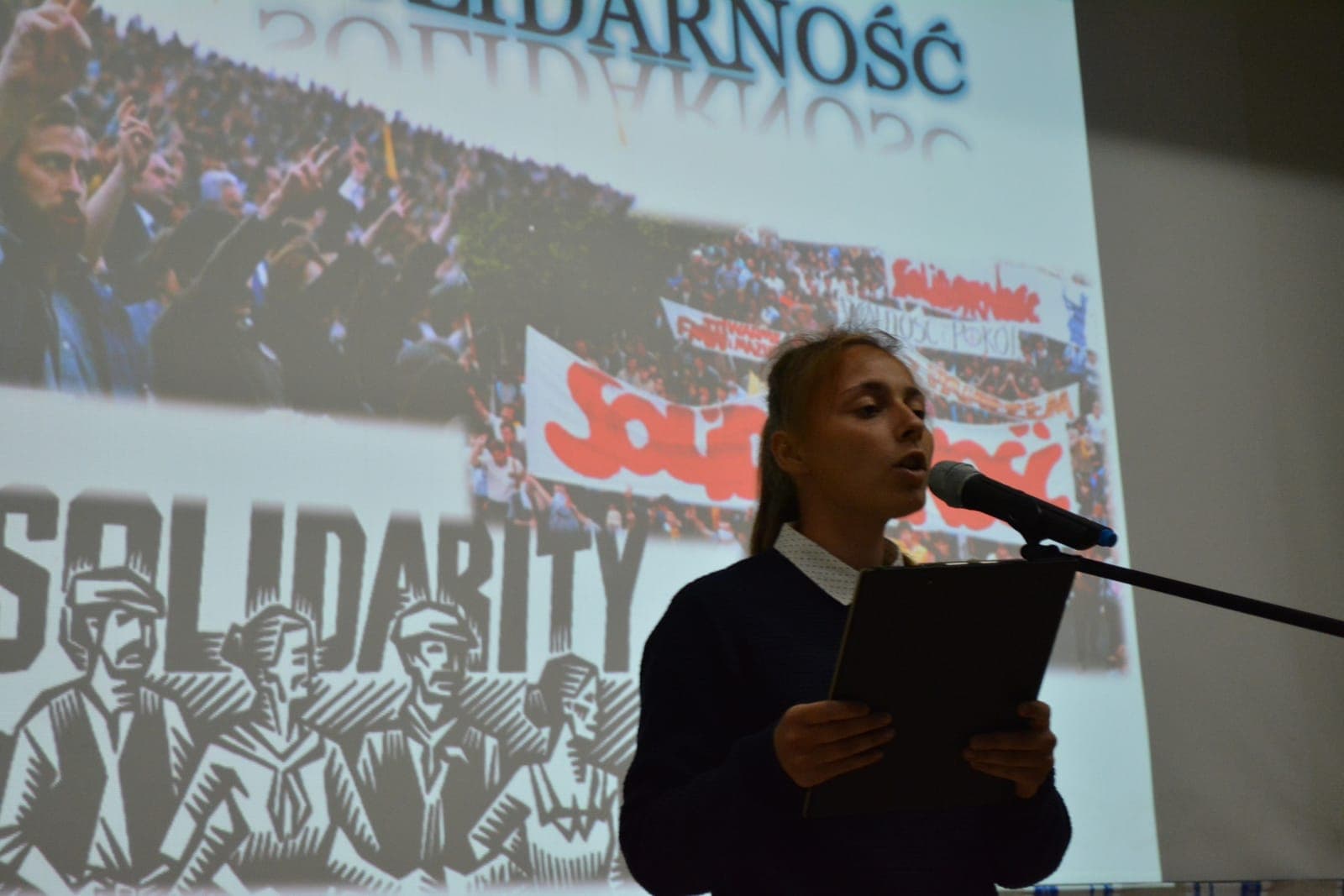 Учениця читає доповідь про «Солідарність» біля мікрофона на тлі проєкції з плакатами Solidarność